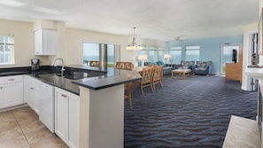 A modern kitchen with a central island, dining area with chairs and a table, a living room with sofas, and large windows offering ocean views.