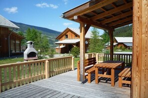 A wooden deck with benches and a stone fireplace, surrounded by cabins with a mountainous backdrop.
