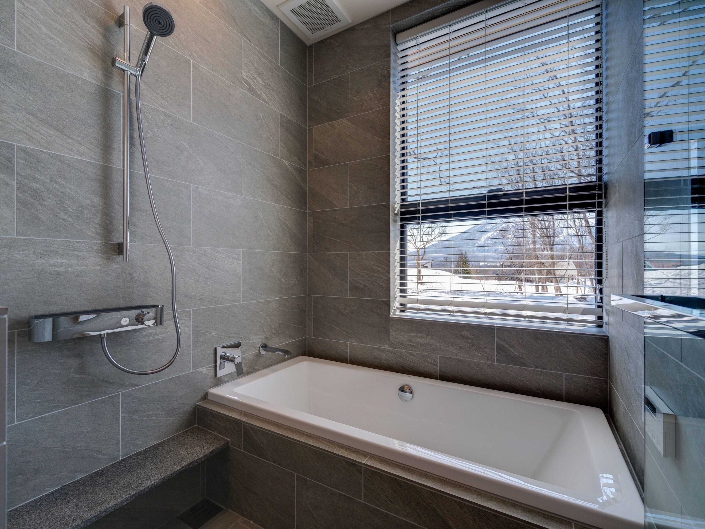 A modern bathroom with a bathtub, shower, and window with blinds.