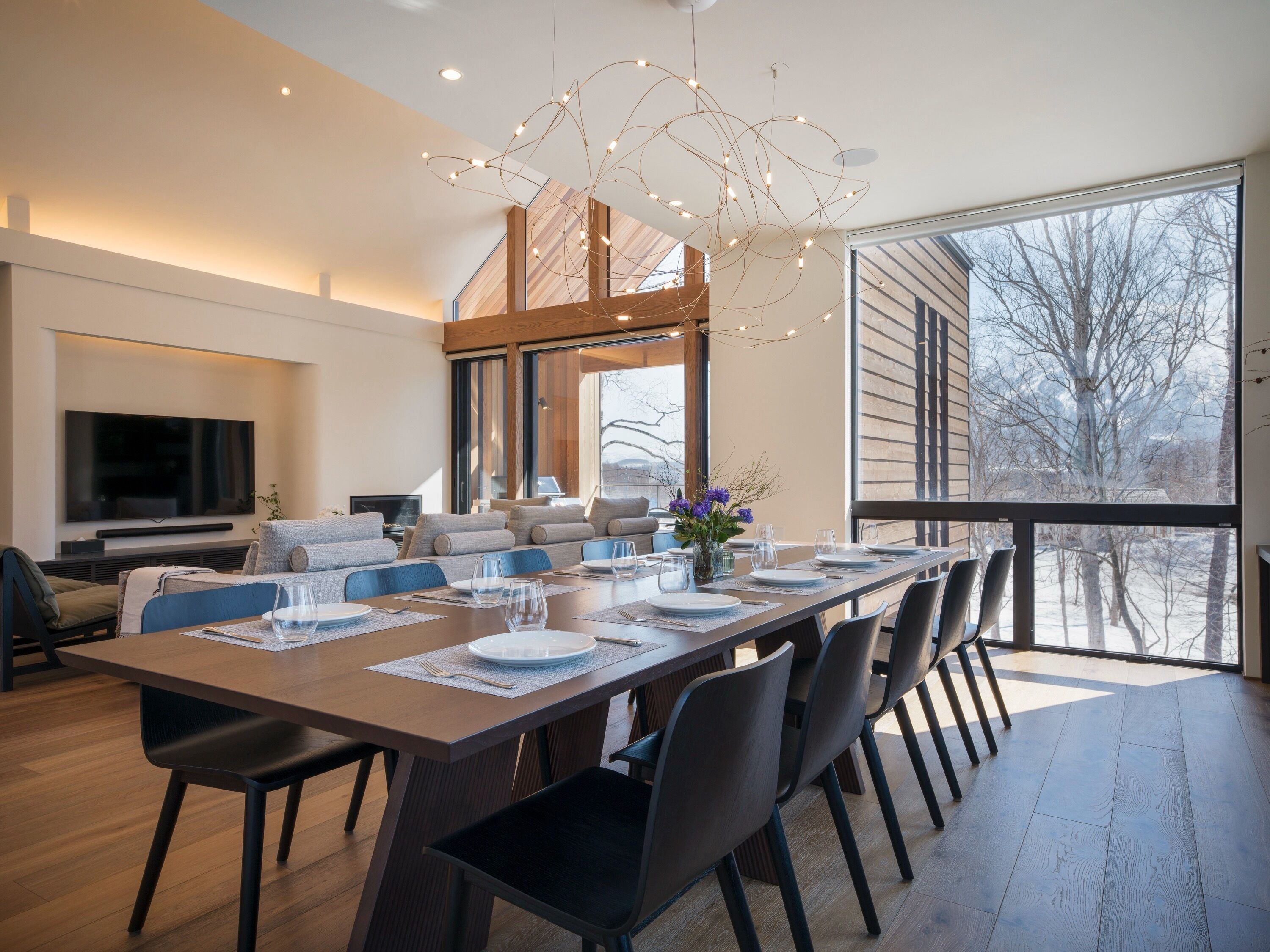 A modern dining area with a long table set for a meal, a television mounted on the wall, and a large window offering a view of the outdoors.