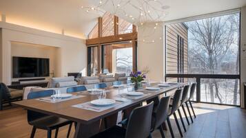 A modern dining area with a long table set for a meal, a television mounted on the wall, and a large window offering a view of the outdoors.
