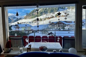 Blick aus dem Fenster auf eine verschneite Berglandschaft mit rotem Geländer und einem Tisch mit Glas und Obstschale.