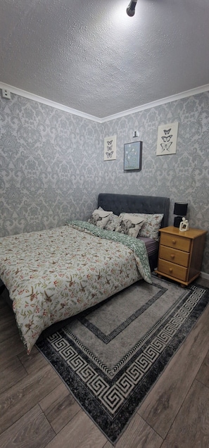 A bedroom with a bed, a nightstand, a lamp, and patterned wallpaper. - Cozy Erith Hideaway - Perfect for Couples (Erith)