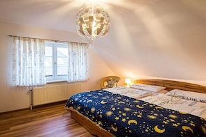 A bedroom with a wooden bed, a night sky-themed bedspread, a window with curtains, a radiator, and a chandelier. - Am Wiesengrund House 14 (Ostseebad Prerow)