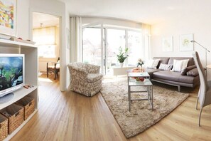 Ein modernes Wohnzimmer mit Sofa, Sessel, Fernseher und einem großen Fenster.