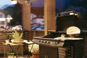Ein Grill mit geöffnetem Deckel, eine Schüssel mit Deckel sowie ein kleiner Tisch mit einer Pflanze.