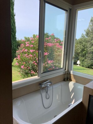A bathroom with a bathtub, a window, and a view of a garden with pink flowers. - Beautiful contemporary architect-designed home with a pool and garden set amid the garrigue (Nîmes)