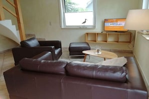 Ein Wohnzimmer mit braunem Ledersofa, einem kleinen Tisch mit einem Glas, einem Fernseher auf einem Holzständer und einer Treppe.