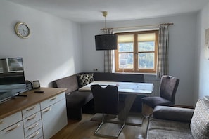 Ein modernes Wohnzimmer mit Fernseher, Sofa, Esstisch und einem Fenster mit Vorhängen.