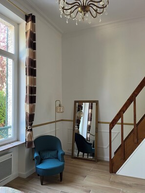A room with a blue armchair, a mirror, a chandelier, and a wooden staircase. - Le Cantilien (Chantilly)