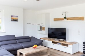 A modern living room with a gray sectional sofa, a wooden coffee table with fruit, a flat-screen TV on a wooden stand, and a white cabinet with a plant. - Holiday home in Dranske with swimming pool (Dranske)