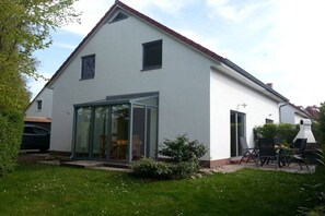 A two-story house with a white exterior, a glass door entrance, and a small garden area with a lawn and a few plants. - Holiday home with 1 bedroom (Ostseebad Rerik)
