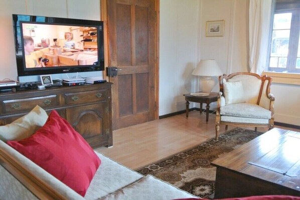 Une pièce comprenant une télévision, un meuble en bois, un canapé avec des coussins rouges, un fauteuil, une lampe et un tapis à motifs.