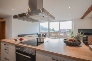 A modern kitchen with an island, built-in oven, stainless steel range hood, and a large window offering a view of the outdoors. - 100 m² Guesthouse Sonne (Bichlbach)