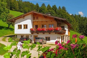 Eine zweistöckige Chalet mit Balkon, umgeben von viel Grün und parkenden Autos.