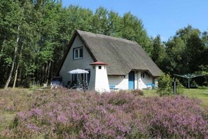 Ein traditionelles Haus mit Strohdach, einem kleinen turmartigen Aufbau und einem Garten mit violettem Heidekraut.