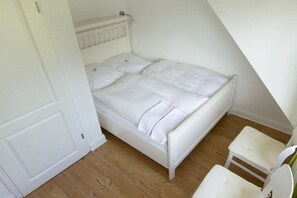 Ein weißes Bett in einem Raum mit schrägem Dach, ein kleiner weißer Stuhl und eine Tür.