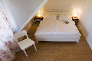 Ein Zimmer mit einem Einzelbett, schrägen Decken, weißer Bettwäsche, zwei Nachttischlampen, einem Sessel und einem Vorhang.