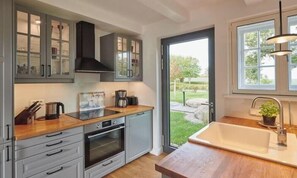 A kitchen with grey cabinets, a wooden countertop, a built-in oven, a sink, and a view of a garden through a glass door. - Holiday home with 4 bedrooms (Friederikenhof)