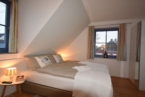 Ein Schlafzimmer mit einem Bett, einer Nachttischlampe, einem Fenster mit Vorhängen und Blick auf Gebäude.