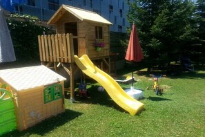 Ein Spielhäuschen aus Holz mit einer Rutsche, einer grünen Spielanlage und einem roten Sonnenschirm.