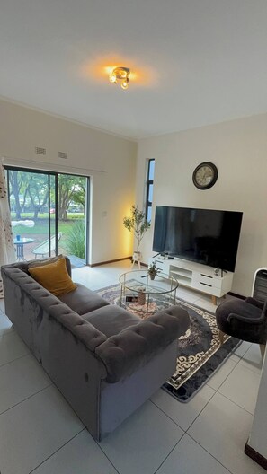 Sala de estar con sofá, televisor, reloj y puerta corrediza de vidrio hacia un patio.
