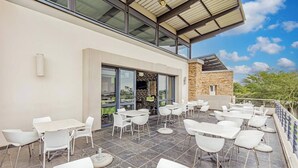 An outdoor patio with white tables and chairs, a stone wall, and a glass door entrance. - Apartment in Northriding (Johannesburg)