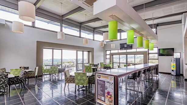 A modern restaurant with high ceilings, large windows, and a mix of green and white pendant lights. - Apartment in Northriding (Johannesburg)