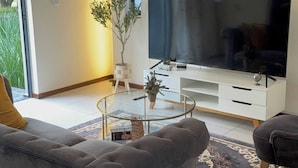 A modern living room with a sofa, a glass coffee table, a flat-screen TV, a clock, and a potted plant. - Apartment in Northriding (Johannesburg)