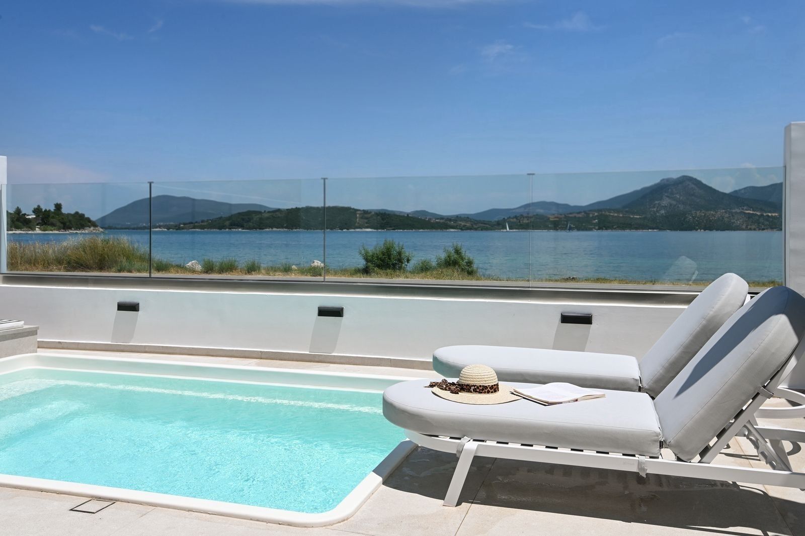 A pool area with a view of a lake and mountains, a lounge chair, and a small table with a hat and book.