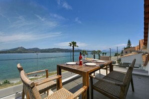 Una mesa de madera con platos y una botella, sillas de mimbre y una vista de un lago con montañas al fondo.