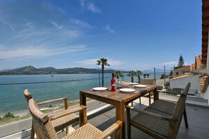 Una mesa de madera con platos y una botella, sillas de mimbre y una vista de un lago con montañas al fondo.