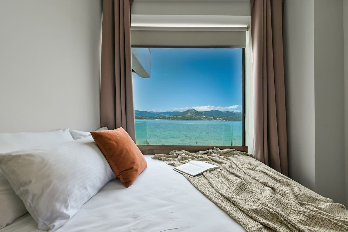 A bed with white linens, a brown pillow, and a knitted blanket. A window reveals a clear sky and a distant mountain range.