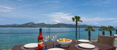 Une table en bois, dressée avec des assiettes, une bouteille d’Aperol et un saladier de fruits, donnant sur une étendue d’eau avec des montagnes en arrière-plan.