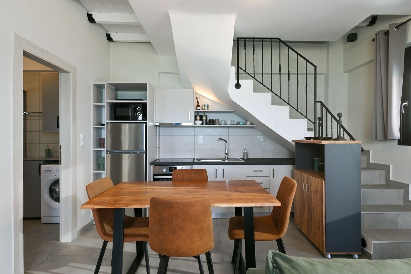 A modern kitchen with a built-in oven, microwave, and sink. There is a dining area with a wooden table and chairs. A staircase with a metal railing leads to an upper level.