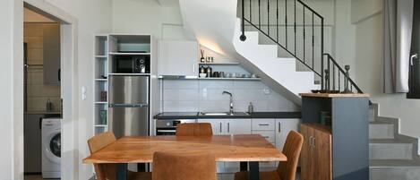 A modern kitchen with a built-in oven, microwave, and sink. There is a dining area with a wooden table and chairs. A staircase with a metal railing leads to an upper level.