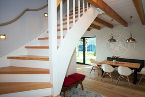 Ein modernes Interieur mit einer Holztreppe, einem Essbereich mit rundem Tisch und Stühlen sowie einem großen Fenster.