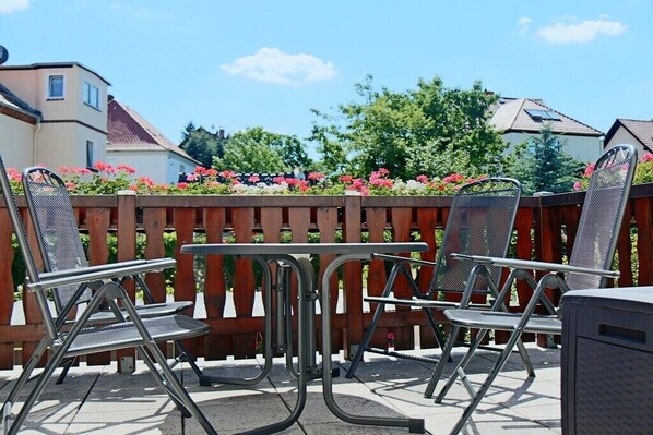 Une terrasse extérieure avec des chaises et une table en métal, entourée d’une clôture en bois et de plantes en fleurs.