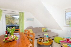 Ein gemütliches Wohnzimmer mit einer Couch, einem Esstisch mit Obstkorb, einem kleinen runden Tisch mit Fernbedienung und einem Fenster mit Vorhängen.