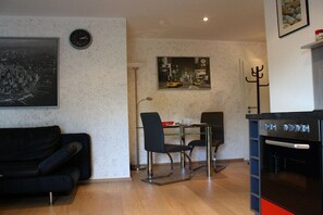Ein Wohnzimmer mit einem schwarzen Sofa, einem Essbereich mit Tisch und Stühlen, einer Uhr und einem Wandbild.