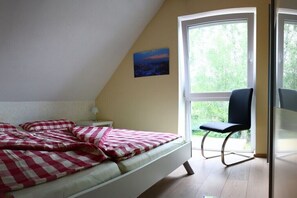 Ein Schlafzimmer mit Bett, Stuhl, Nachttisch, Lampe und einem Fenster mit Blick auf Grünfläche.
