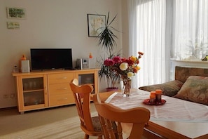 Ein Wohnzimmer mit Fernseher, Holzschrank, Esstisch, Stühlen, einer Vase mit Blumen und einem Sofa.