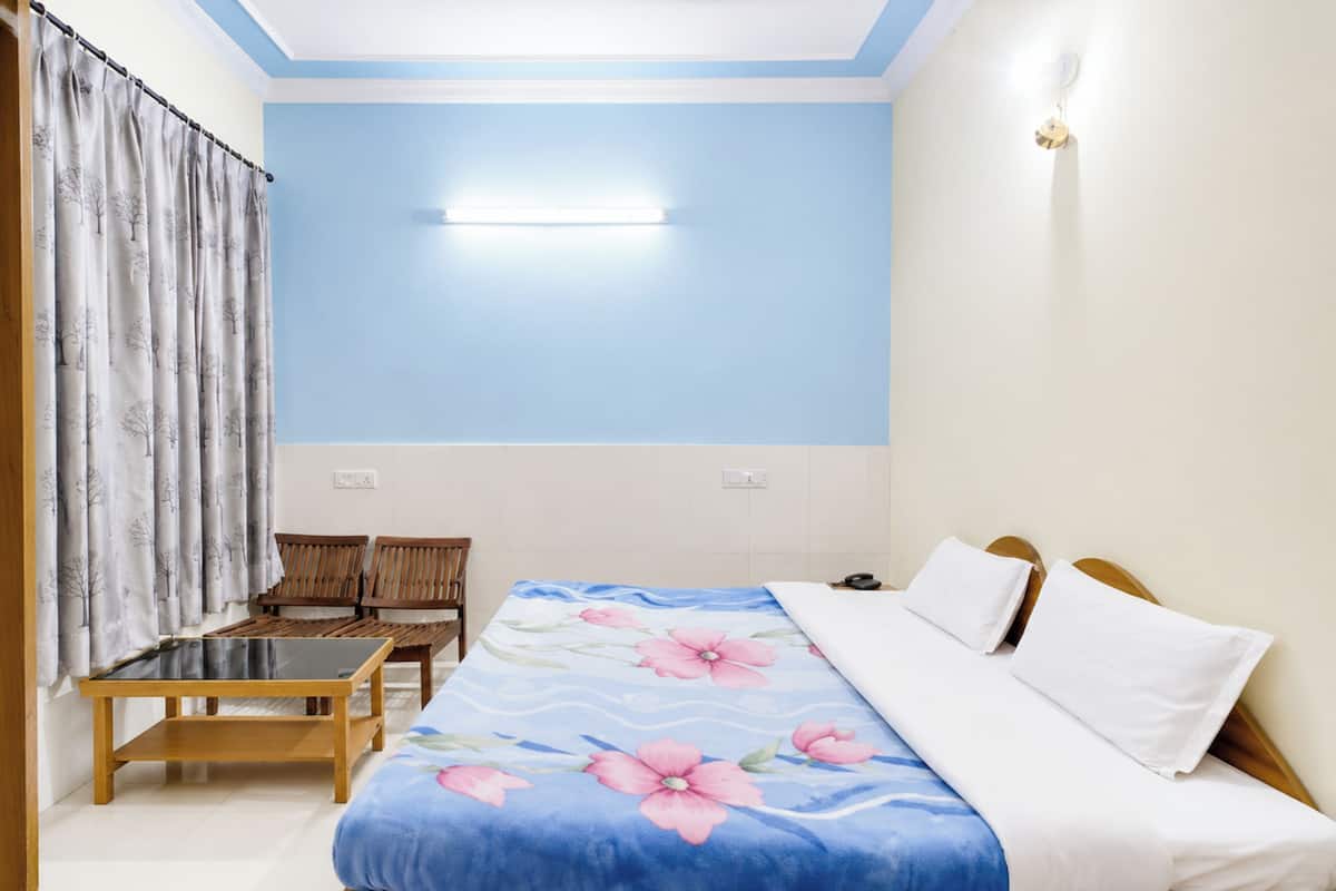 Quarto com cama, duas cadeiras de madeira, uma mesa pequena e luminária na parede.