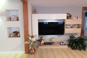 Ein modernes Wohnzimmer mit einem Flachbildfernseher an einer weißen Wand, Holzregalen mit Dekoration, einer Vase mit Blumen und einem Topfpflanzen.