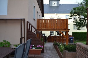 Eine Holzterrasse mit einer Treppe, die zu einem Balkon im zweiten Stock führt, umgeben von einem Pflasterweg und Blumenbeeten.