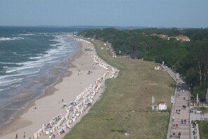 Ein Strand mit einer langen Reihe aus Sonnenschirmen und Liegestühlen, einer Grünfläche und einem Pfad entlang des Wassers.