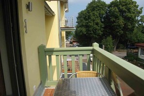 Ein Balkon mit einem Metalltisch und einem Korbstuhl, mit Blick auf eine Straße mit Bäumen und parkenden Autos.