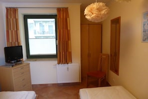 Ein Schlafzimmer mit Bett, Stuhl, Kommode, Fernseher, Fenster mit Vorhängen und Heizkörper.