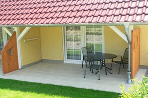 Eine überdachte Terrasse mit Tisch und Stühlen, eine Holztür und ein Fenster mit Blick ins Grüne.