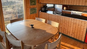 Interior de madera con una mesa redonda para comer, sillas, una lámpara colgante, una zona de cocina con estufa y una ventana con vistas a un paisaje natural.